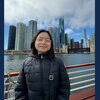 A woman in a black coat and glasses stands in front of a building skyline.