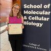 A blonde woman in a pink top and glasses smiles while holding a certificate.