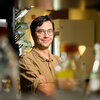 A man in a tan shirt and glasses stands in front of lab beakers.