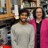 A woman in a pink blazer and a man in a grey sweater stand in a laboratory.