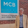 Man in blue shirt and beige jacket stands in atrium in front of sign that says MCB, School of Molecular & Cellular Biology
