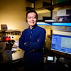 Wei Qin in blue shirt stands in front of computer screen and microscope
