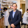 Wearing a blue blazer, Nash Kalsotra stands in a biochemistry lab between two benches while two graduate students behind him