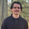Man with brown hair and mustache stands outside wearing dark colored sweater