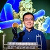 Wei Qin holds a petri dish in front of projection screen showing bacteria. Photo by Fred Zwicky.