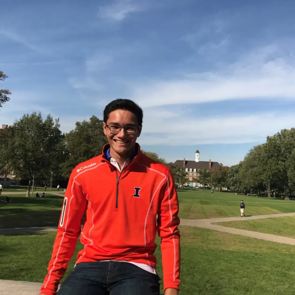 Michael Kraft sits in an orange U of I quarter zip sweatshirt on the Quad, with Foellinger Auditorium in the background.