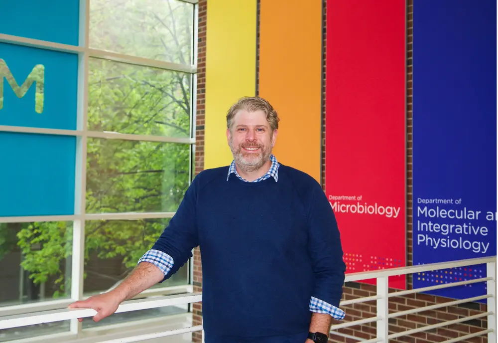 A man in a blue sweater smiles in front of a brightly-colored background
