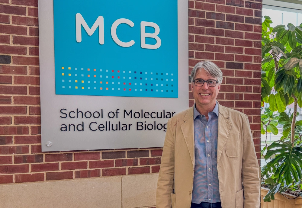 Man in blue shirt and beige jacket stands in atrium in front of sign that says MCB, School of Molecular & Cellular Biology