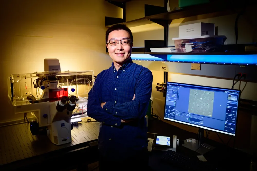 Wei Qin in blue shirt stands in front of computer screen and microscope