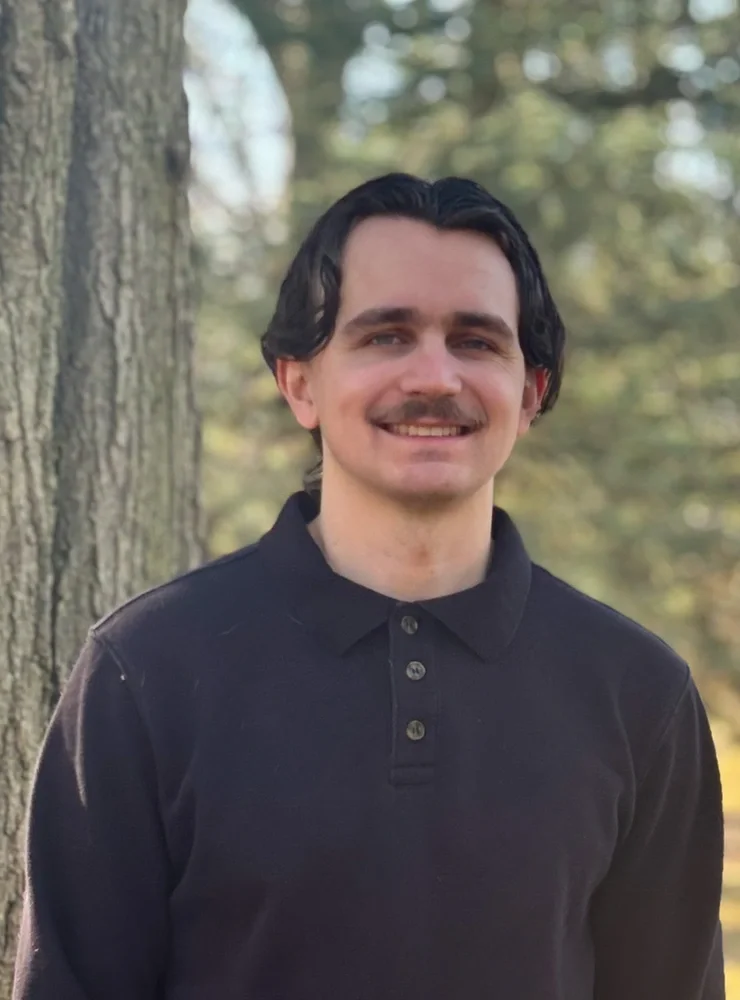 Man with brown hair and mustache stands outside wearing dark colored sweater