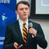 Brendan Rattin stands in a suit and tie holding a microphone in one hand and a remote in the other.