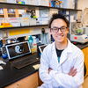 Yiquan Wang sits smiling in a white lab coat with his arms crossed next to a computer displaying his artwork in the Wu Lab.