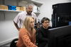 Drs. Kristen Flatt and Yiwu Zheng sit at a computer monitor. They are looking at the monitor, with Dr. Satish Nair standing behind them looking at the screen as well.