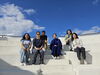 Six students sit on a white staircase in front of a blue sky.