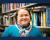 A smiling woman standing in front of a bookshelf wearing a blue shirt and scarf