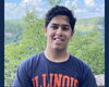 A man in a blue and orange shirt standing in front of a tree line.