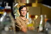 A man in a tan shirt and glasses stands in front of lab beakers.