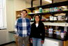 A man in a blue checkered shirt and a woman in a navy sweater stand in front of a lab bench.