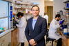 Wearing a blue blazer, Nash Kalsotra stands in a biochemistry lab between two benches while two graduate students behind him