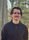 Man with brown hair and mustache stands outside wearing dark colored sweater